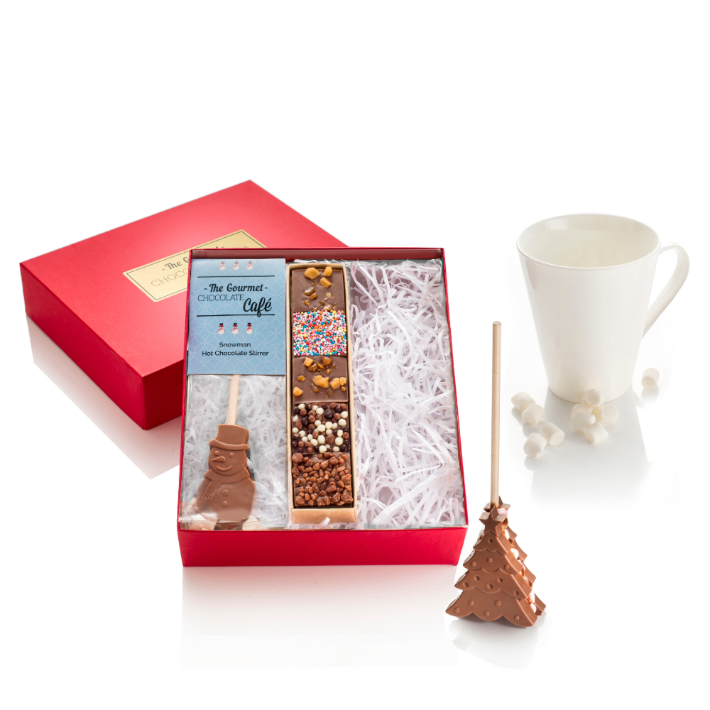 Red gift box with a snowman hot chocolate stick and chocolate mendiants. A christmas tree hot chocolate stick is presented next to a mug with marshmallows.